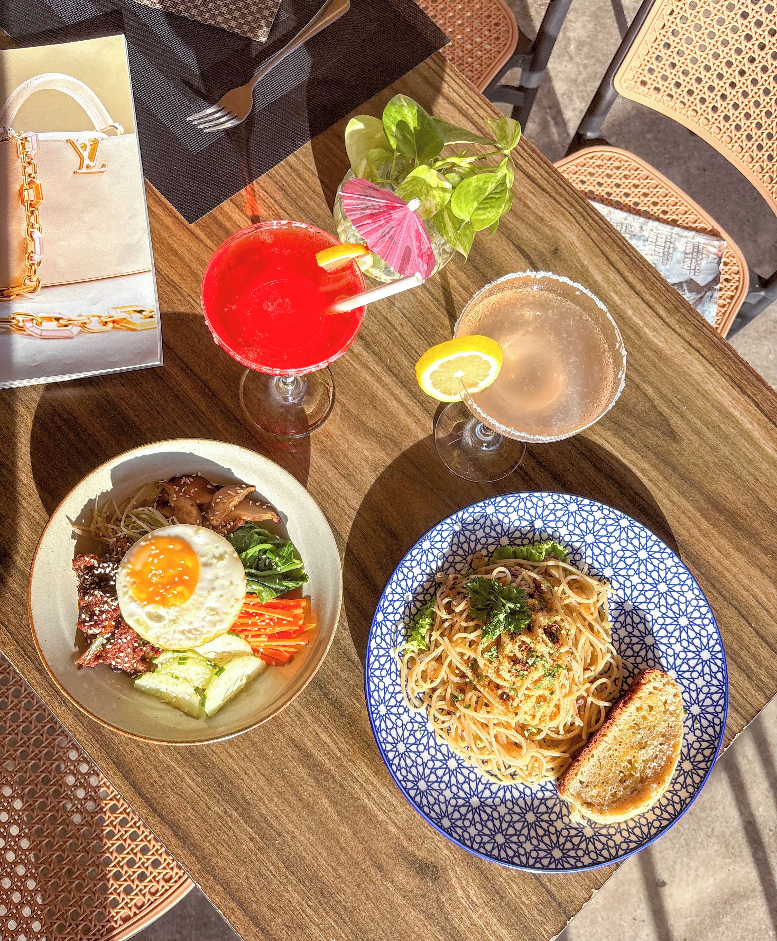 A table with a variety of dishes including noodles, a fried egg, and a bowl of soup, along with two drinks and a handbag.