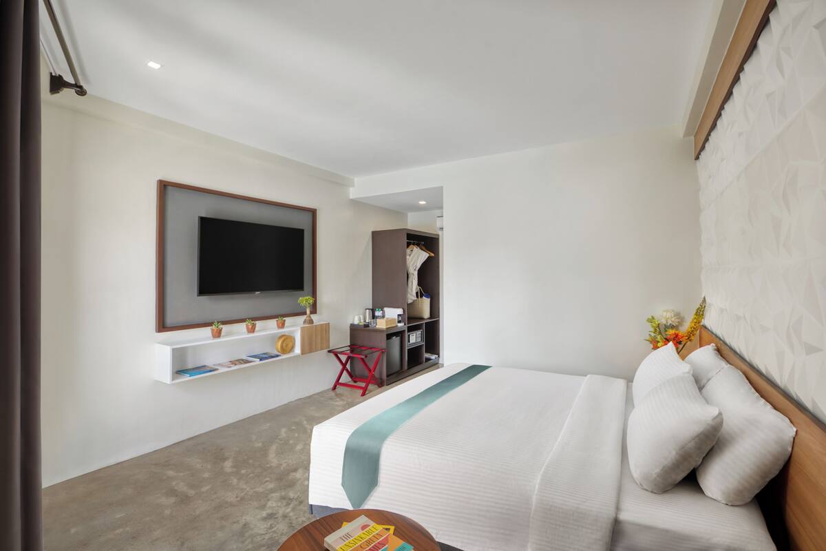 Ein modernes Hotelzimmer mit einem großen Bett, einem an der Wand montierten Fernseher, einem kleinen Schreibtisch und einem Kleiderschrank mit Kleidung.