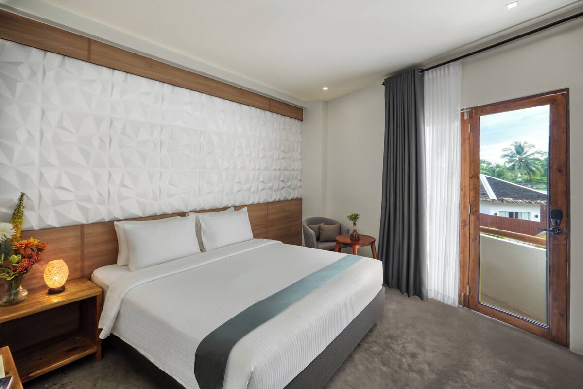 Ein modernes Hotelzimmer mit einem großen Bett, einem Holzkopfende, einem Nachttisch mit Lampe, einem kleinen Tisch mit Blumenvase und Blick ins Freie durch eine Schiebetür.