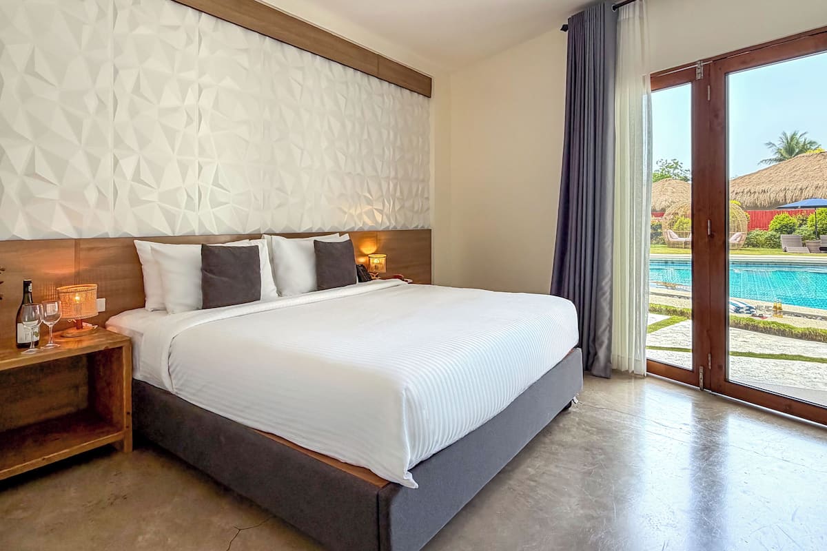 Ein Hotelzimmer mit einem großen Bett, einem Nachttisch mit Lampe und Blick auf einen Pool.