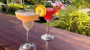 Zwei Cocktails auf einem Holztisch mit Blick auf einen tropischen Garten und strohgedeckte Gebäude.