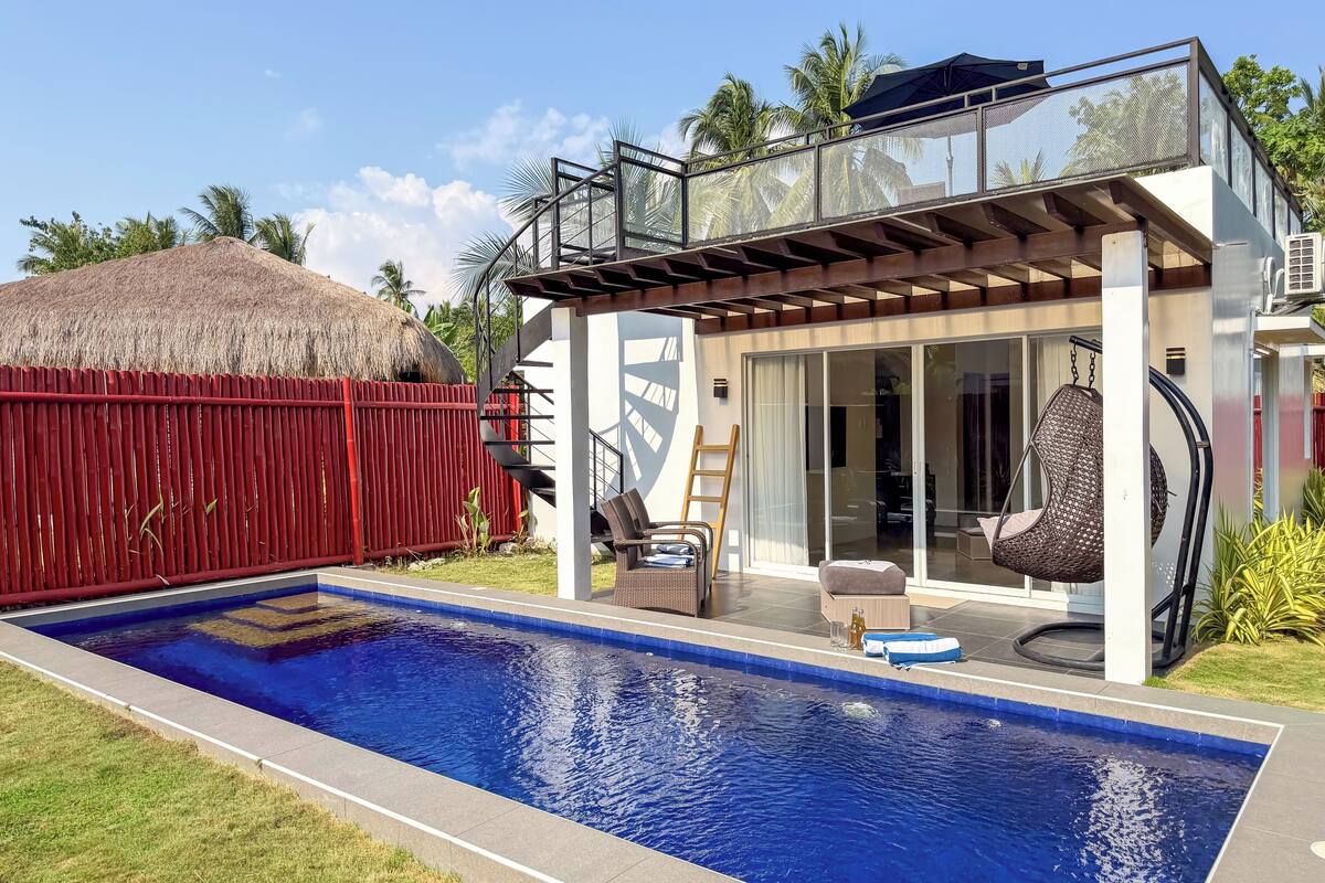 Ein modernes Haus mit Pool, rotem Zaun und einem Reetdach-Anbau.