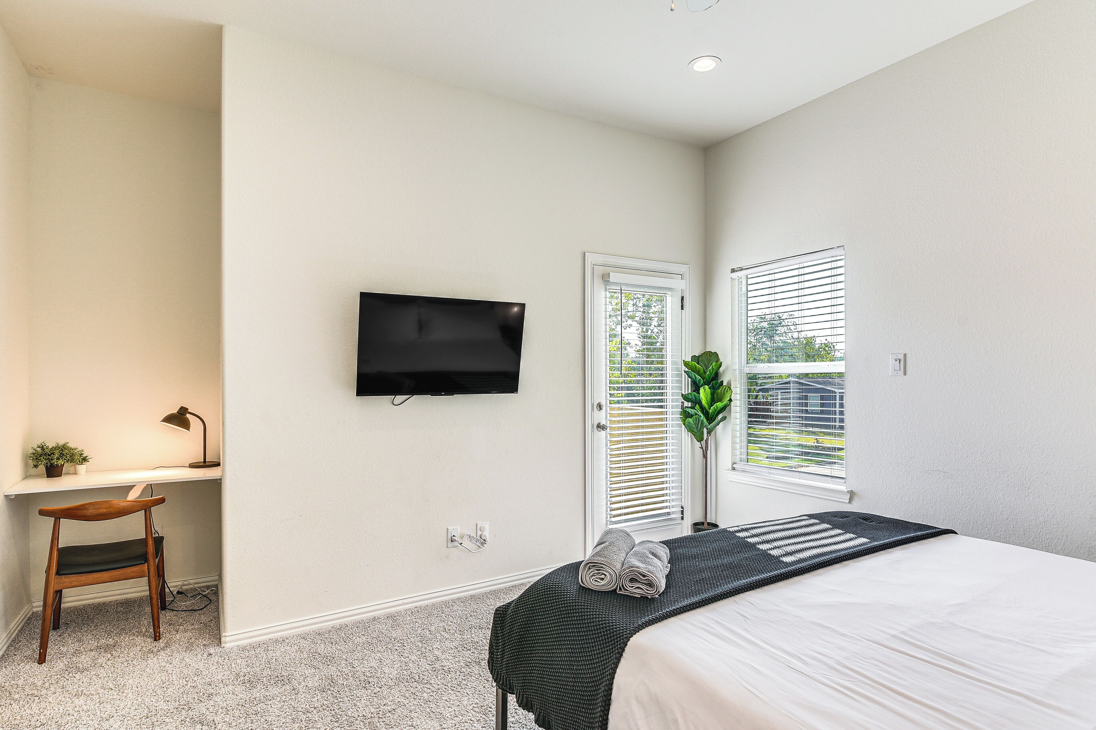 A bedroom with a bed, a desk, a chair, a TV, and a window.