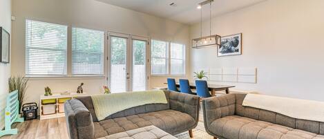 A modern living room with a sectional sofa, a dining area, and large windows with blinds.