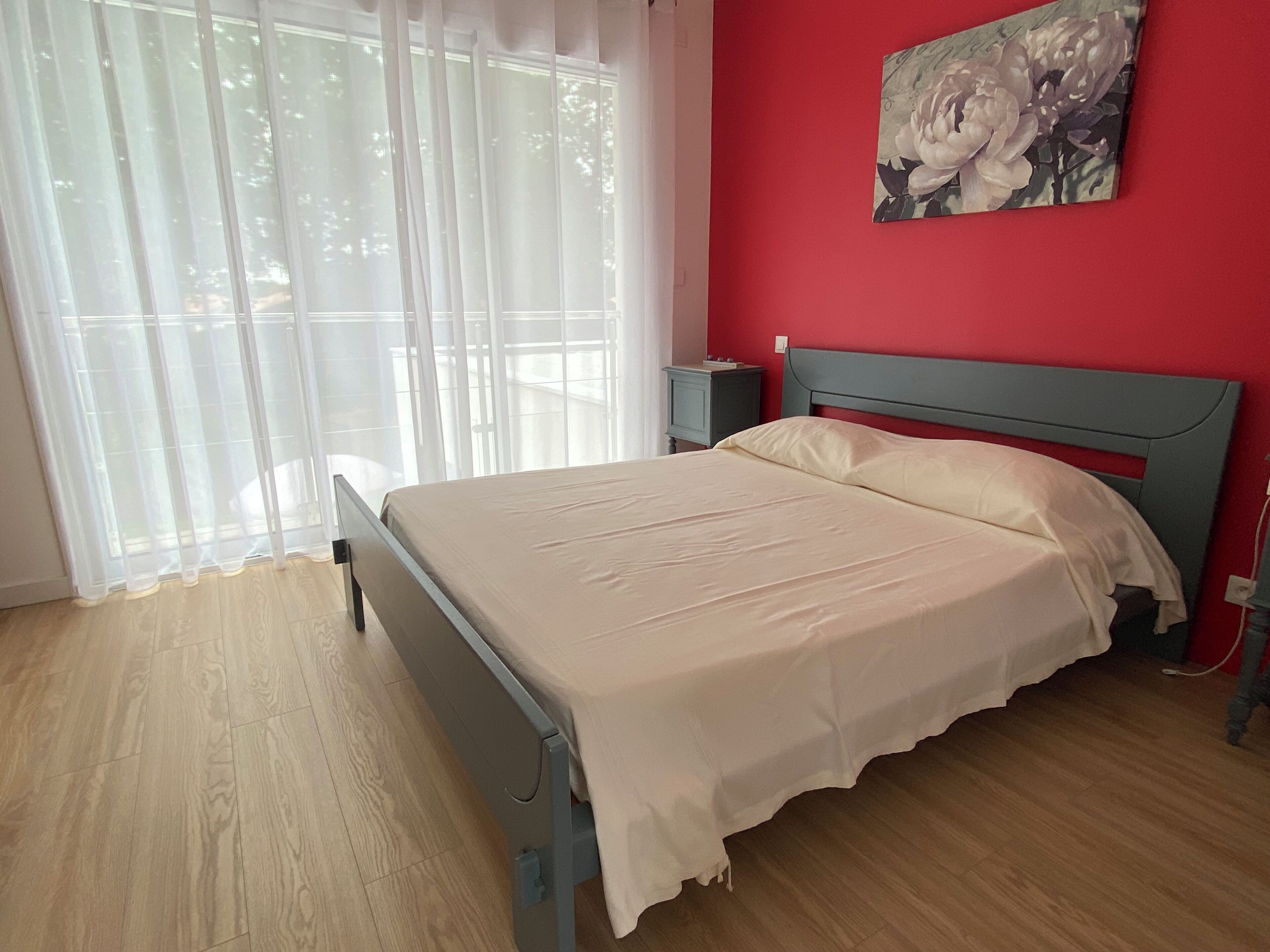 Une chambre à coucher avec un lit, une table de chevet, un mur rouge et une fenêtre ornée de rideaux légers.