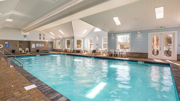 An indoor swimming pool with clear water, surrounded by white chairs and tables, and a brick pathway leading to the pool.