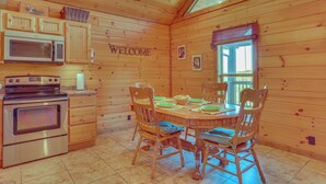A wooden cabin interior with a kitchen area, a dining table set for four, and a 'WELCOME' sign on the wall.