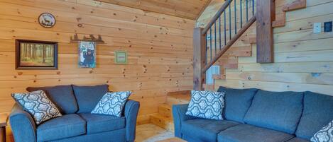 A log cabin interior with two blue sofas, a wooden coffee table, a staircase, and a welcome sign.