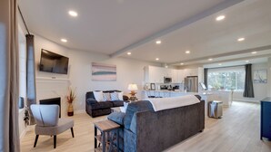 A modern living room with a sofa, armchair, and a small table. There is a fireplace, a television mounted on the wall, and a kitchen area in the background. - Balconies w/ Scenic River Views: Hammond Townhome! (Hammond)