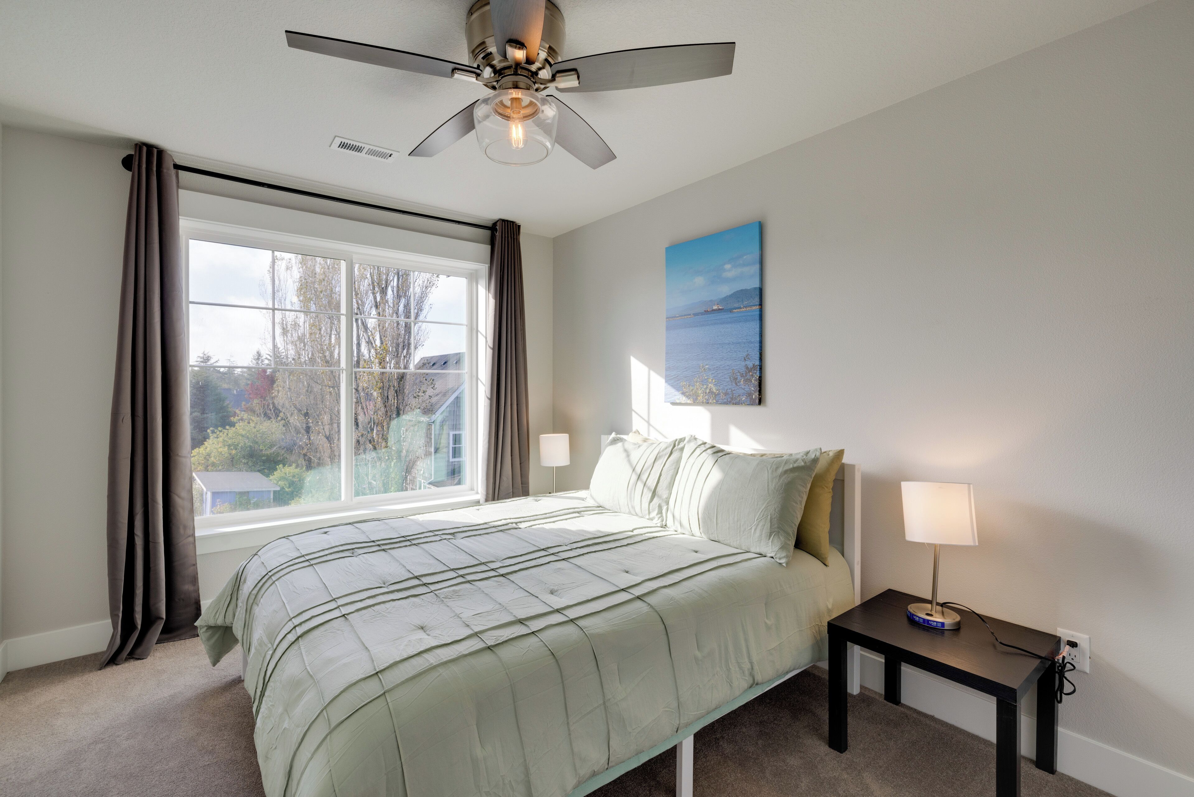 A bedroom with a bed, a nightstand, a ceiling fan, a window with curtains, and a painting on the wall.