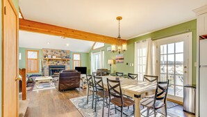 A spacious living room with a fireplace, a dining area with a wooden table and chairs, and a kitchen with a refrigerator and a window. - Beach Access, Private Dock: Arrowhead Lake Cabin (Pocono Lake)