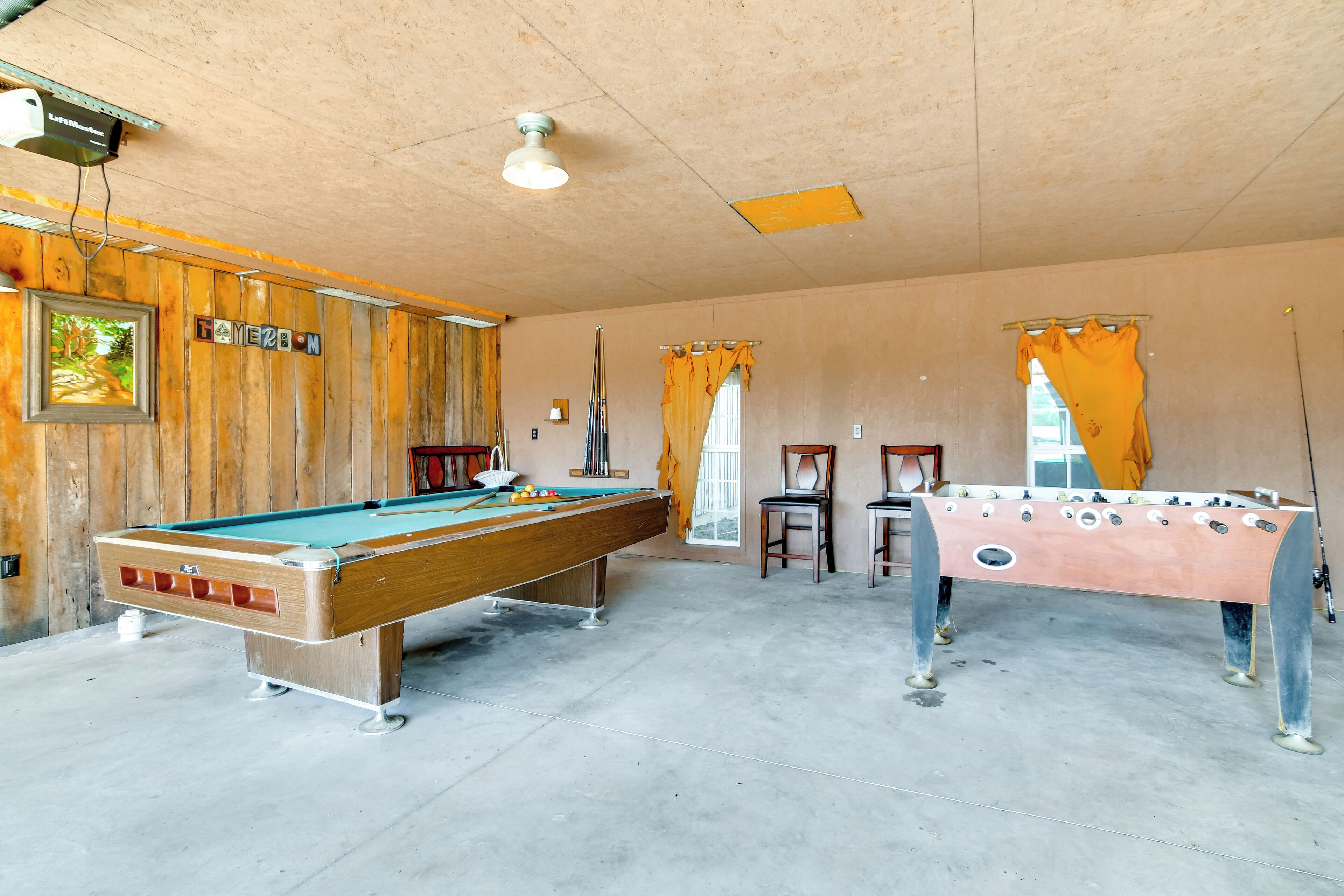 A pool table with a wooden finish, a foosball table, and a billiards table in a room with wooden paneling and a concrete floor.