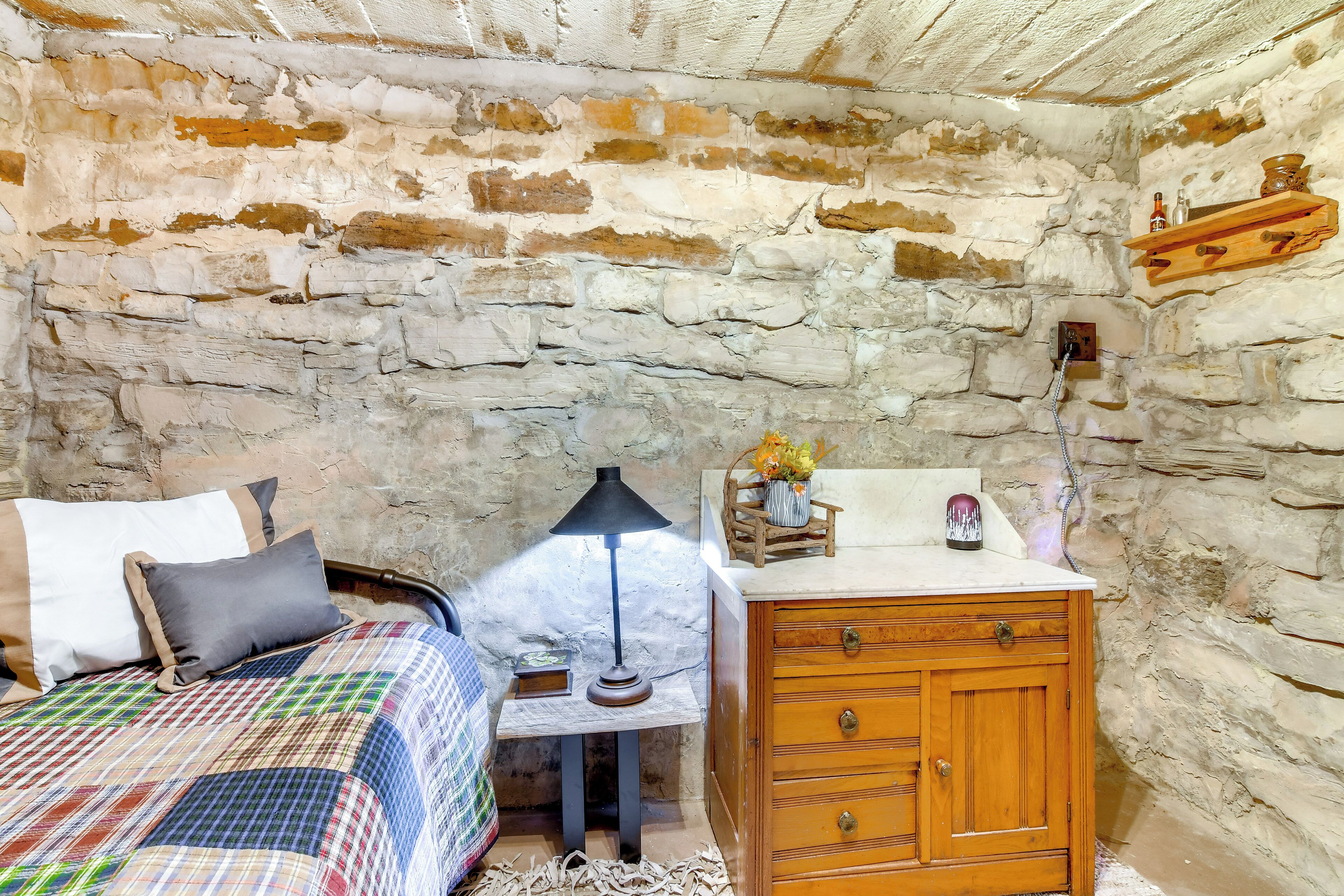 A rustic room with exposed stone walls, a wooden cabinet, a bed with a plaid bedspread, and a small table with a lamp and decorative items.