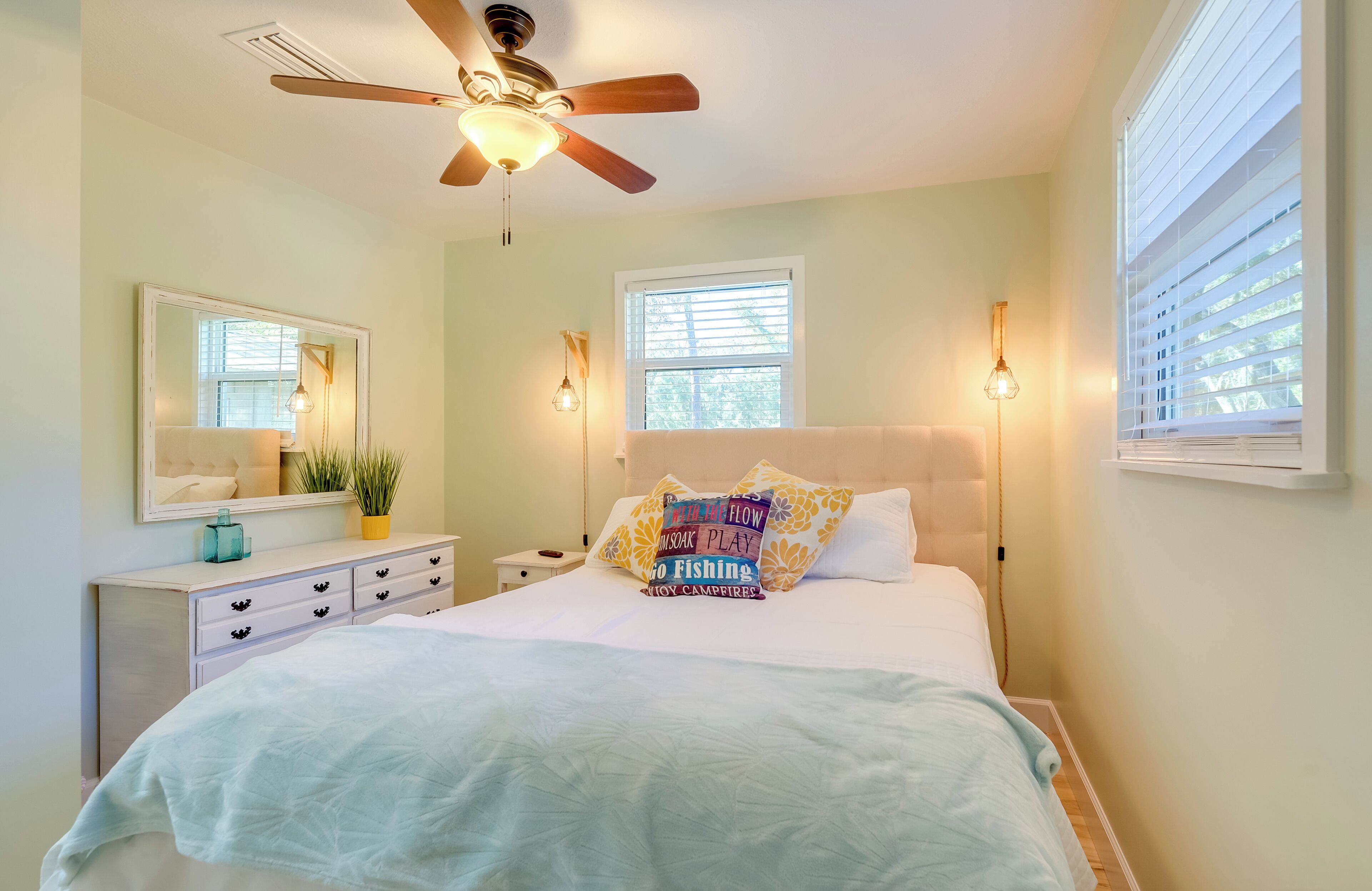 A bedroom with a bed, ceiling fan, bedside lamps, a nightstand, a dresser, and a window with blinds.