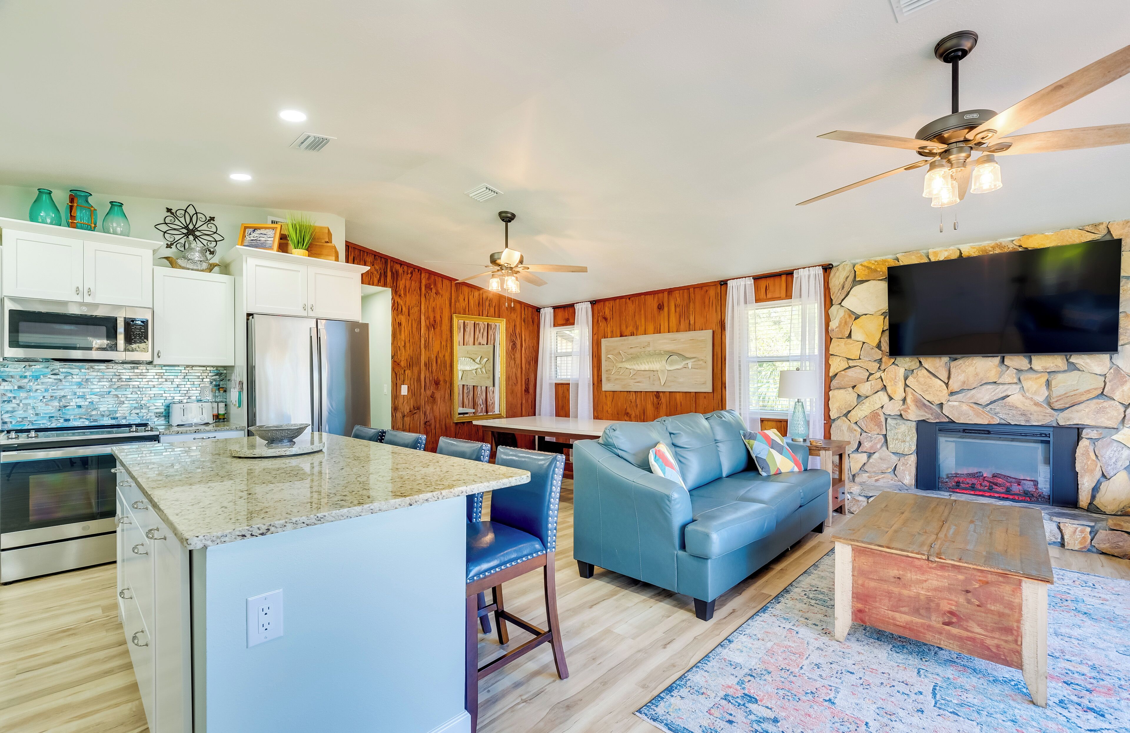 A modern kitchen with granite countertops, a built-in oven, and a refrigerator. There is a dining area with a blue leather couch and a stone fireplace.