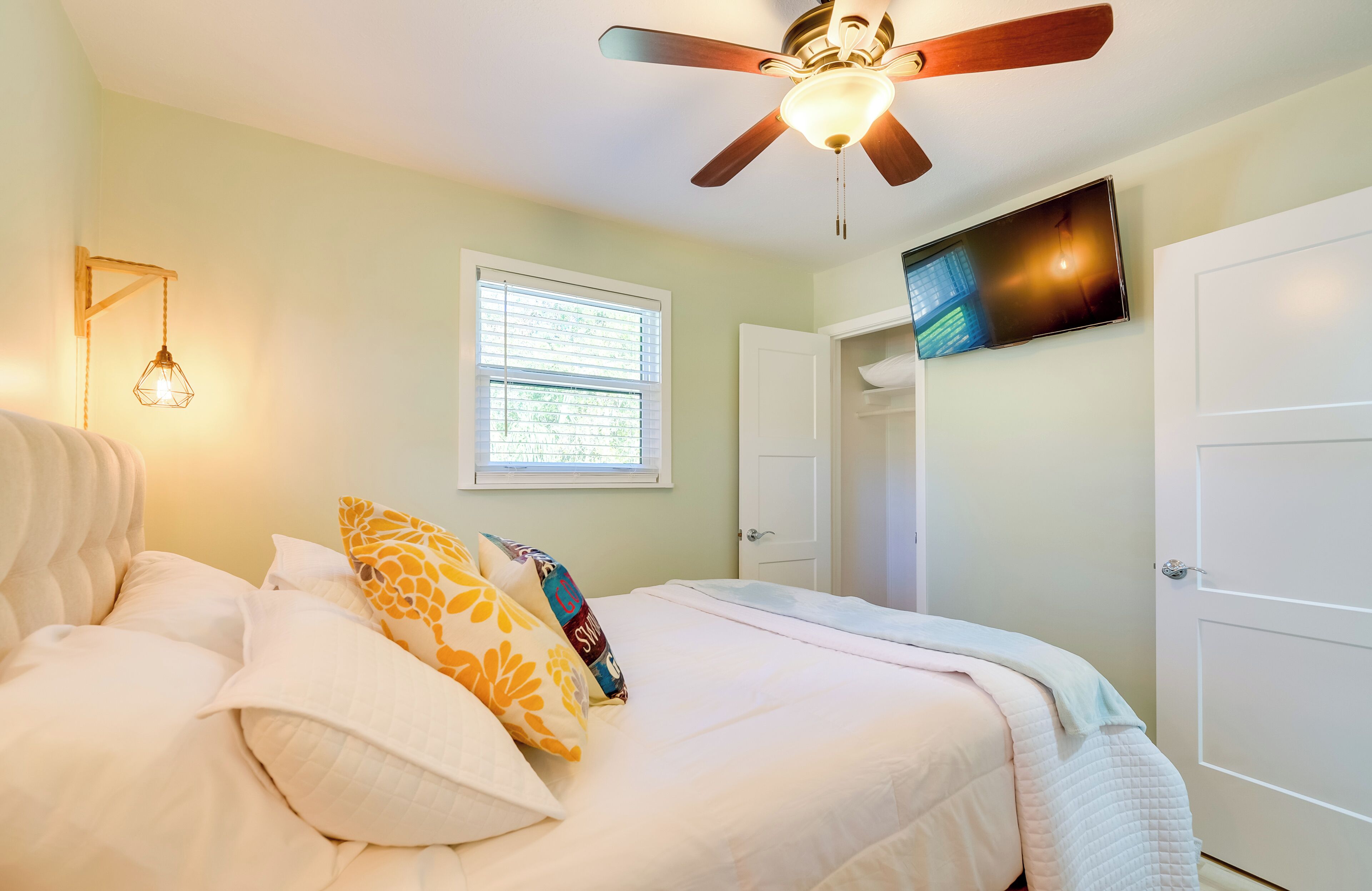 A bedroom with a bed, a ceiling fan, a window, a TV, and a door.