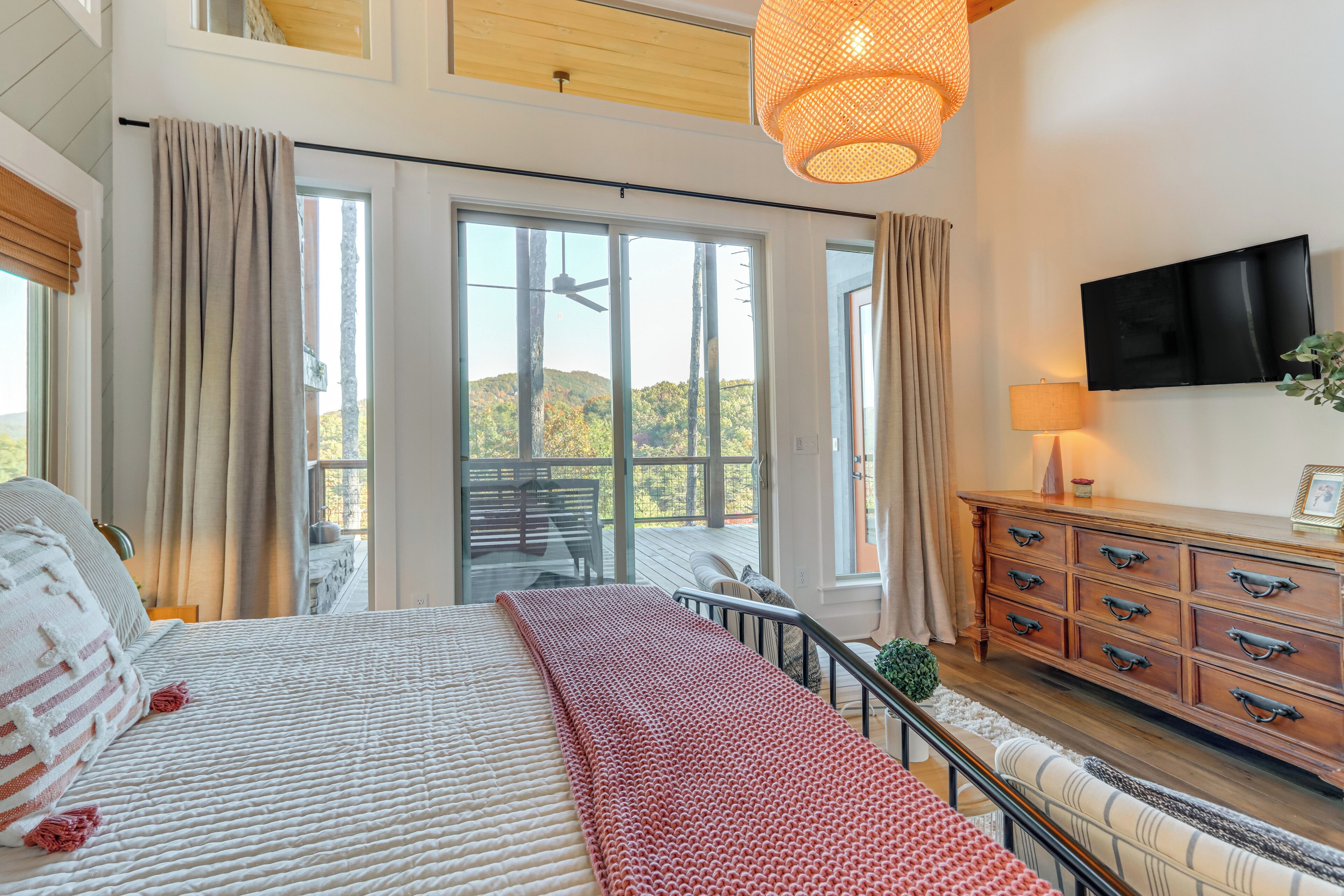 A bedroom with a large bed, a wooden dresser, a TV, and a balcony with a view of trees.