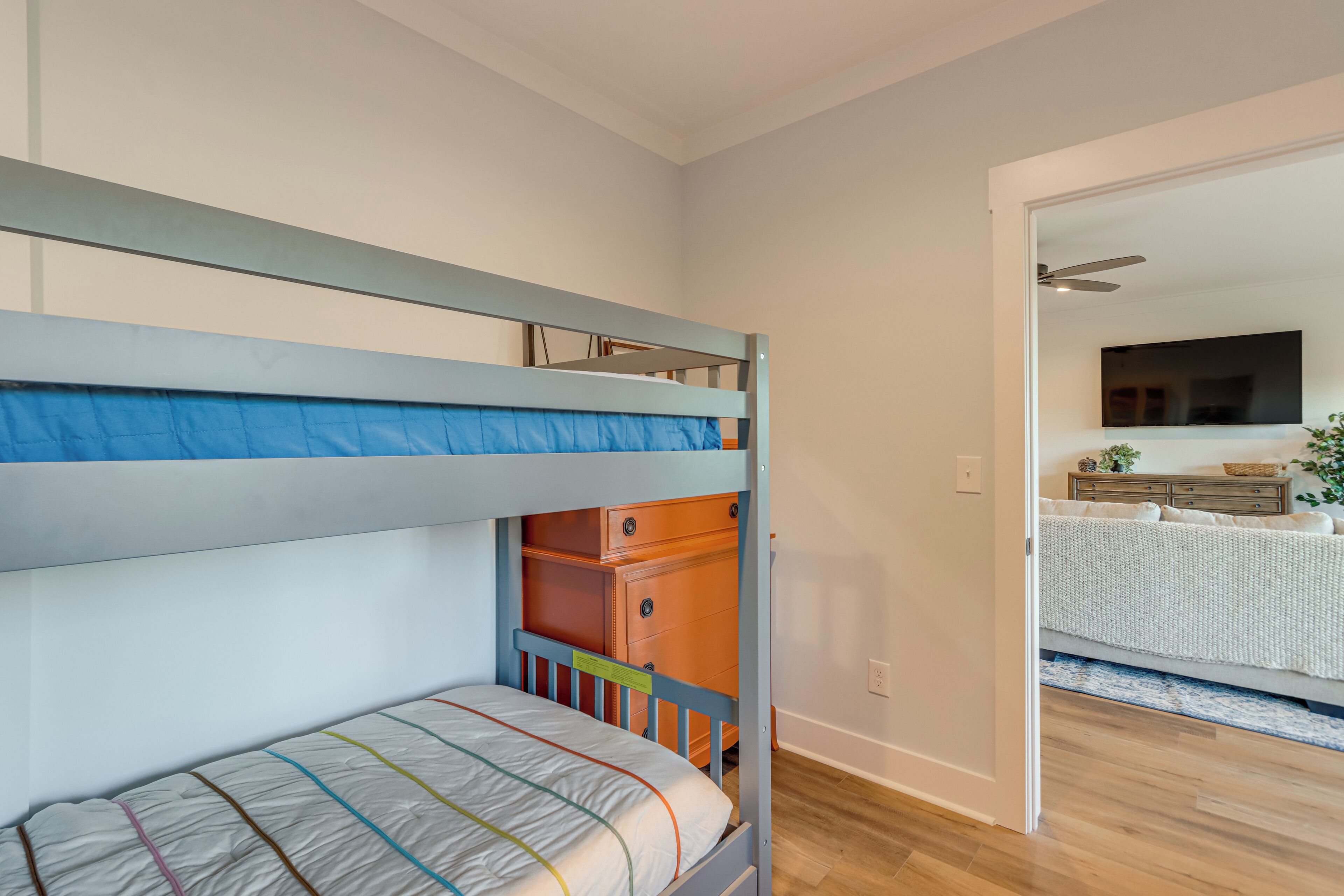 A bunk bed with a lower bunk and a drawer below, a wooden dresser, and a doorway leading to a room with a bed and a TV.