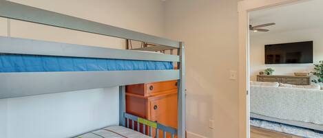 A bunk bed with a lower bunk and a drawer below, a wooden dresser, and a doorway leading to a room with a bed and a TV.