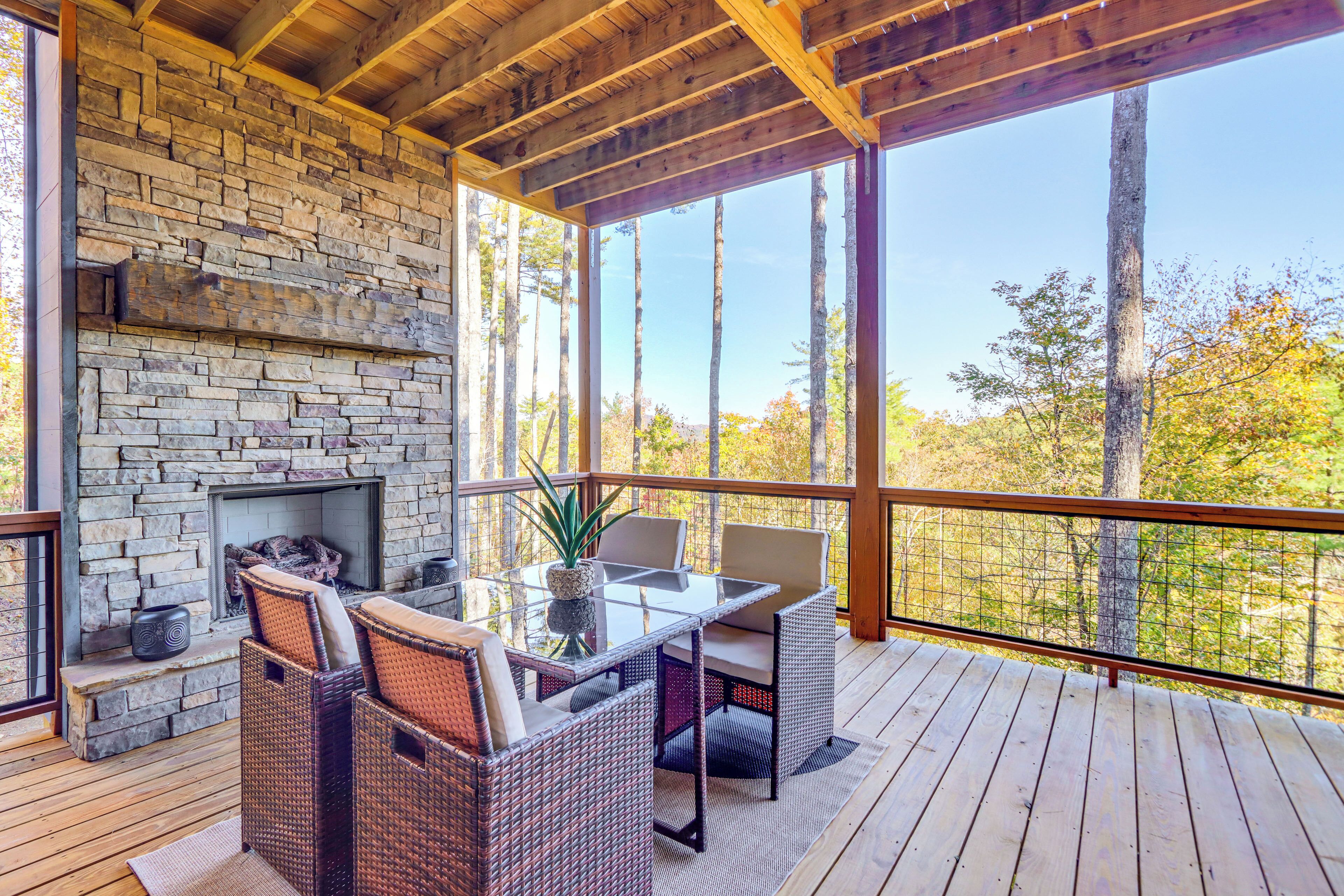 A wooden deck with a stone fireplace, wicker furniture, and a view of trees.