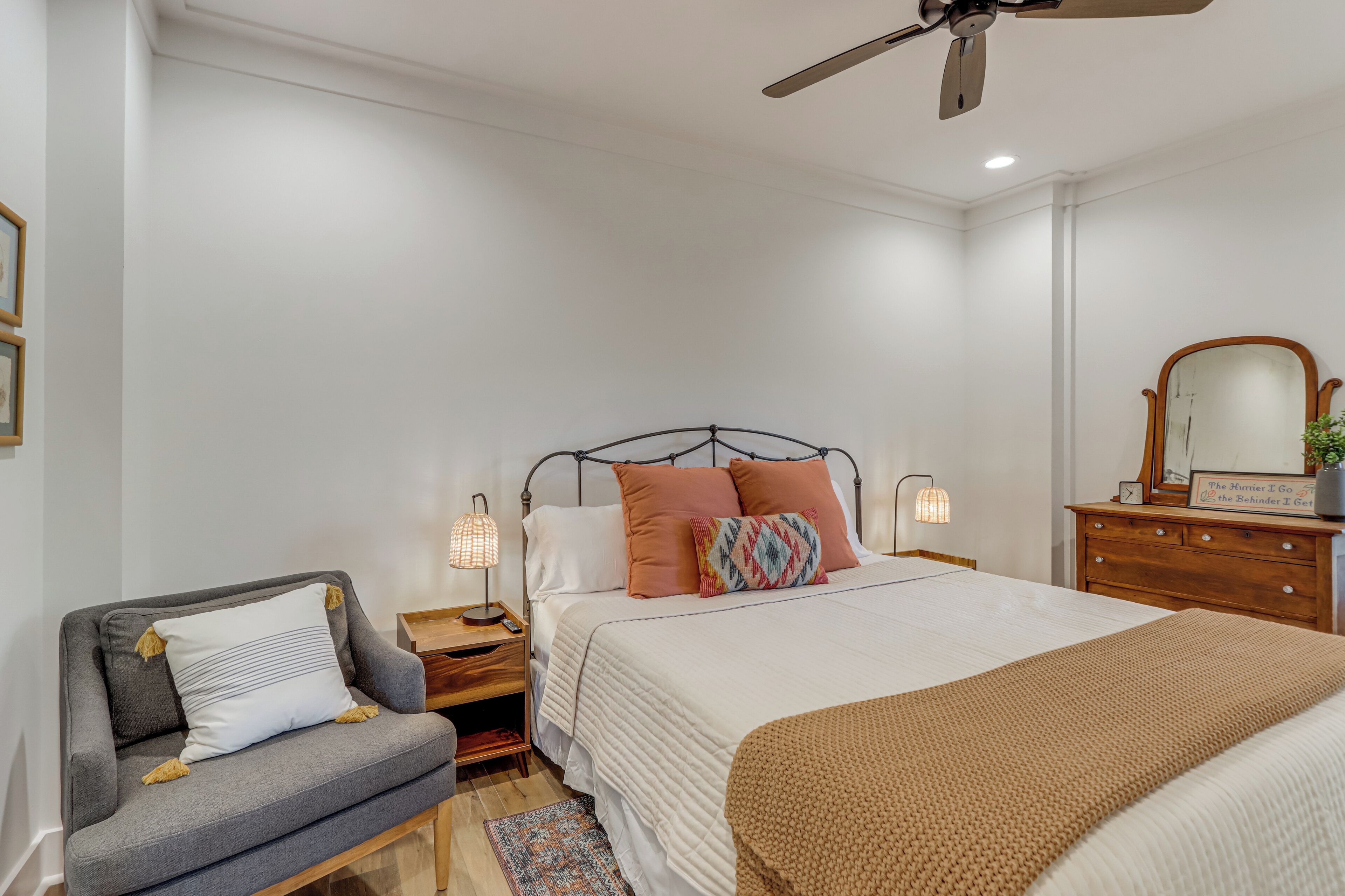 Een slaapkamer met een bed, nachtkastjes, een stoel, een dressoir, een plafondventilator en een spiegel.