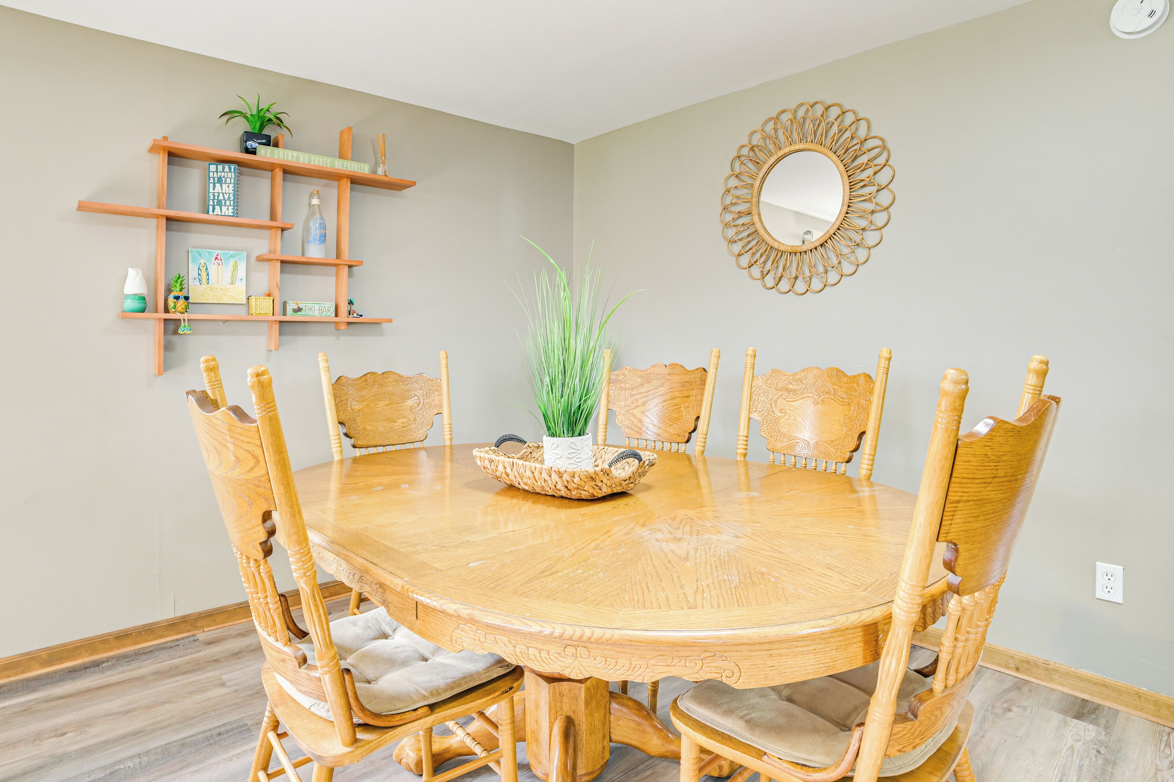 A dining area with a round wooden table surrounded by six chairs, a decorative mirror, and a wooden shelf with various items.