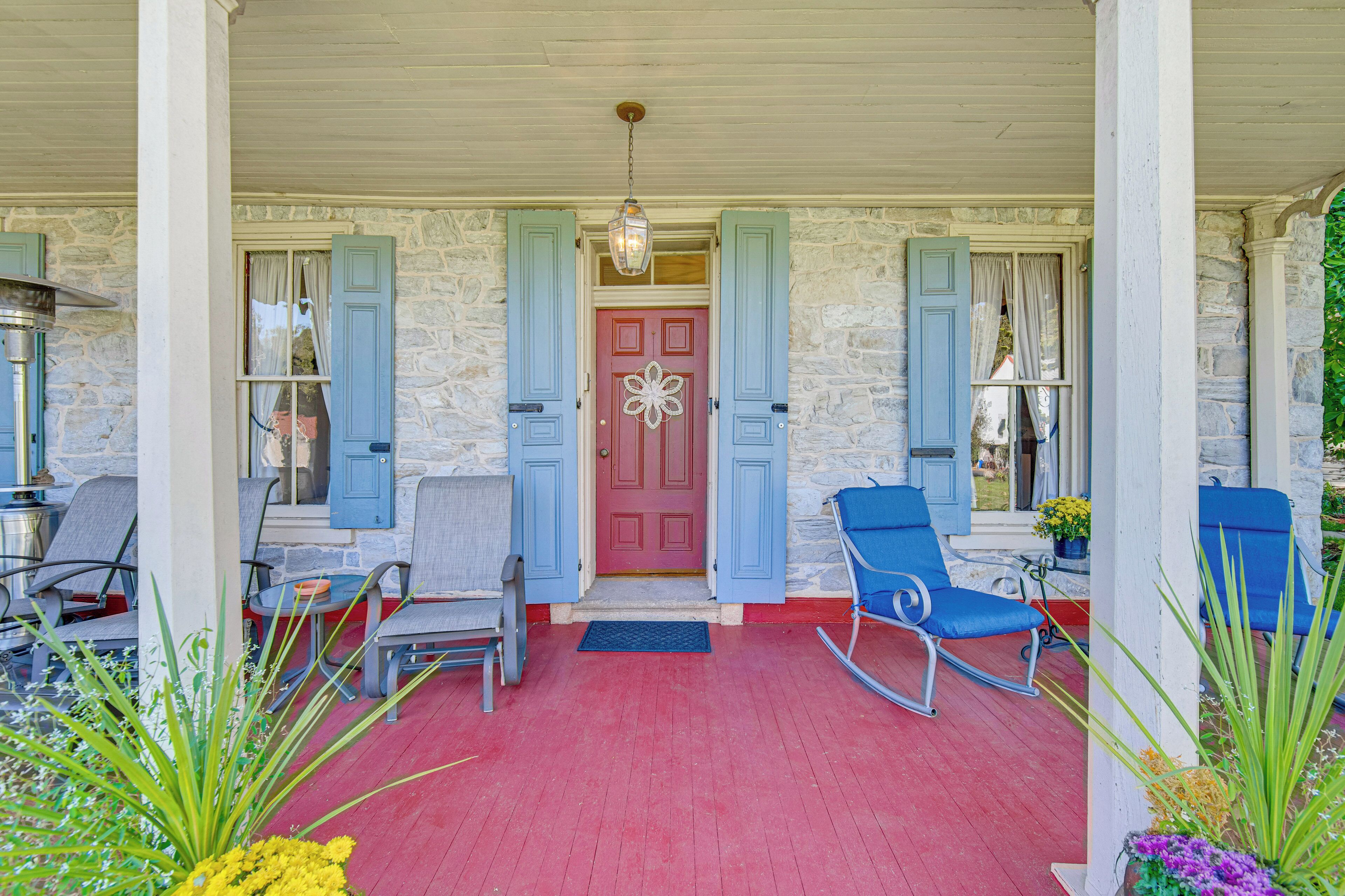A porch with a red floor, stone walls, and blue shutters. There are two rocking chairs and a table with chairs.