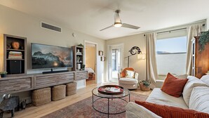 A modern living room with a flat-screen TV, a ceiling fan, and a large window with a view of the sea. - Fully Appointed Upper Floor Condo with Mountain-Canyon-Pool-Golf Course Views (Tucson)
