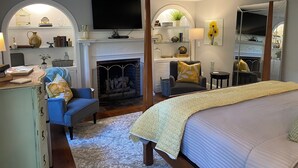 A bedroom with a fireplace, a television, a bed, a blue armchair, and a desk with a lamp. - Inn at Perkins Cove (Ogunquit)