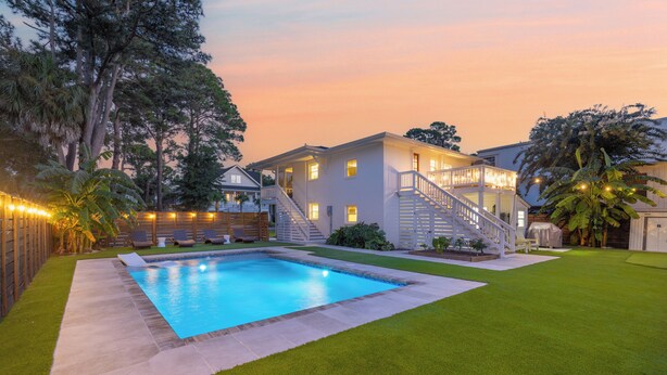 A swimming pool with a wooden deck, a grassy lawn, and a two-story house with a staircase and balcony. - Updated 4th row beach home, private pool, fenced in yard, and putting green! (Hilton Head)