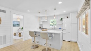 A modern kitchen with white cabinetry, a central island with bar stools, pendant lighting, and a view of the outdoors through a window. - Updated 4th row beach home, private pool, fenced in yard, and putting green! (Hilton Head)