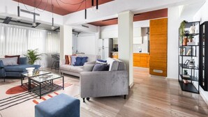 Un soggiorno moderno con divano, poltrone e tavolino. È presente una zona cucina con pensile in legno e scaffale nero con piante.