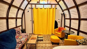 Un interior acogedor con cama, sofá y una pequeña mesa, que destaca por un techo abovedado único y una cortina amarillo brillante.