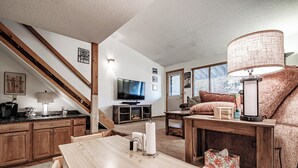 A cozy living room with a wooden staircase, a flat-screen TV, a fireplace, and a comfortable seating area with a lamp. - One bedroom Cascades C-7, sleeping 6, fireplace! (Newry)