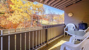 A covered outdoor deck with a view of autumn trees, a wooden railing, and white plastic chairs. - One bedroom Cascades C-7, sleeping 6, fireplace! (Newry)