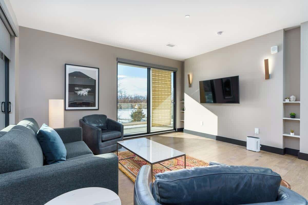 Uma sala de estar moderna com sofá, poltrona e mesa de centro, uma TV fixada na parede e uma grande janela com vista para o exterior.