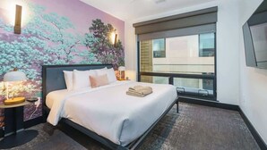 Quarto de hotel moderno com uma cama grande, mesa de cabeceira, TV fixada na parede e janela com vista para o prédio.