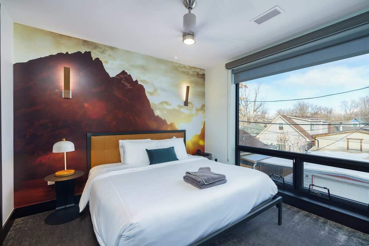 Ein Hotelzimmer mit einem großen Bett, einem Nachttisch mit Lampe, einem Wandbild und einem großen Fenster mit Blick nach außen.