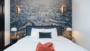 Hotelkamer met een bed, wit beddengoed, een rode handdoek, twee bedlampen en een groot muurschildering van een besneeerd landschap.