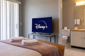 A hotel room with a bed, a television displaying the Disney+ logo, a bathroom with a toilet and sink, and a window with blinds. - 3BR Townhome Steps From Theme Parks Sleeps 6 (Kissimmee)