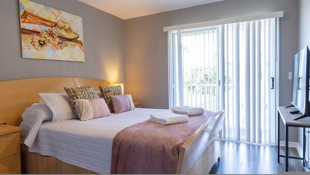 A bedroom with a bed, pillows, a bedside table, a TV, and a window with blinds.