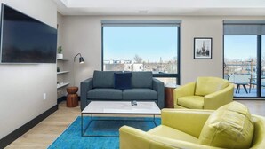 Un salon moderne avec un canapé gris, des fauteuils jaunes, une table basse et de grandes fenêtres donnant sur la ville.