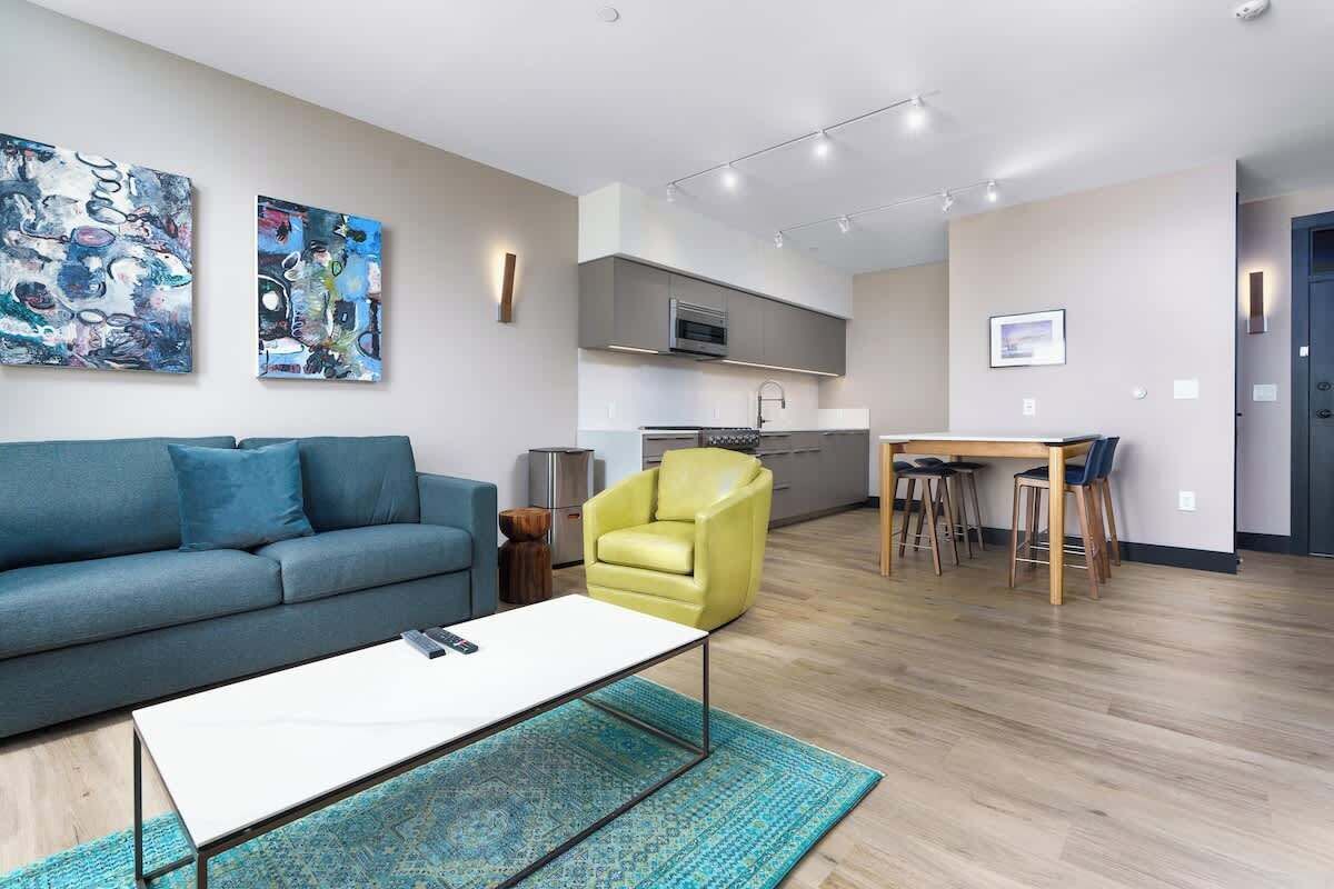 A modern living room with a sofa, coffee table, and a yellow armchair. There is a dining area with a table and chairs. The room has a kitchen in the background.