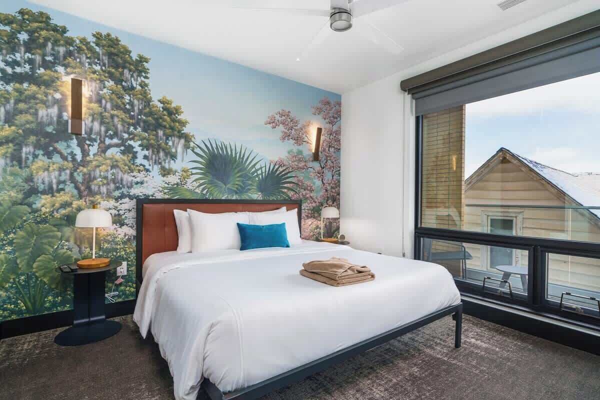 A bedroom with a large bed, a nightstand, a lamp, and a mural of trees and flowers on the wall.