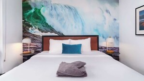 Habitación de hotel con una cama grande, dos lámparas de noche, un almohárga azul y un mural de una ola.