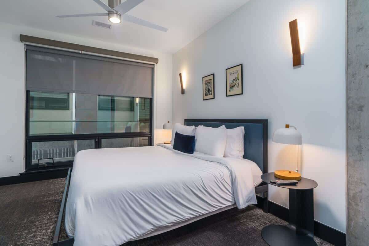 A hotel room with a large bed, a ceiling fan, a window with blinds, a bedside table with a lamp, and framed pictures on the wall.