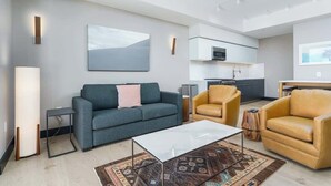 Un salon moderne avec un canapé gris, une table basse et deux fauteuils en cuir. On aperçoit une cuisine avec des meubles blancs et un four à micro-ondes à l’arrière-plan.