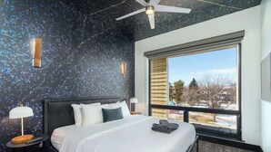 A modern bedroom with a large bed, a ceiling fan, a nightstand, a lamp, and a window with a view of buildings and trees. - Sleek Tennyson St Flat w/ Balcony & Views (Denver)