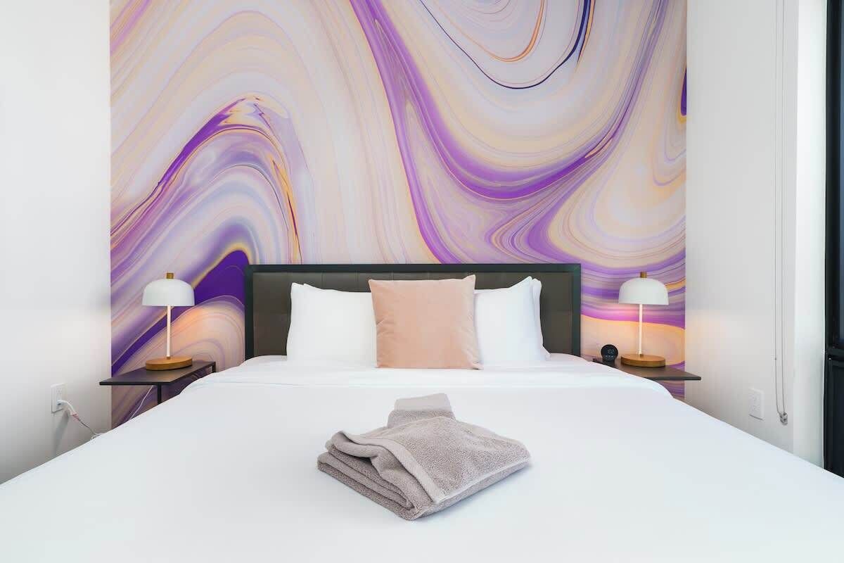 A modern hotel room with a large bed, two bedside tables with lamps, and a vibrant abstract wall mural.