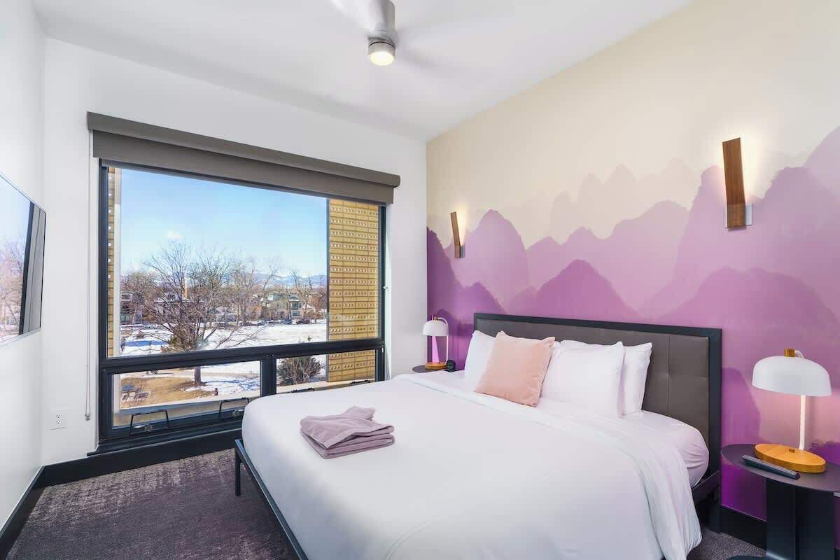 Quarto de hotel moderno com uma janela grande, cama com roupa branca, mesa de cabeceira com abajur e um mural de montanhas na parede.